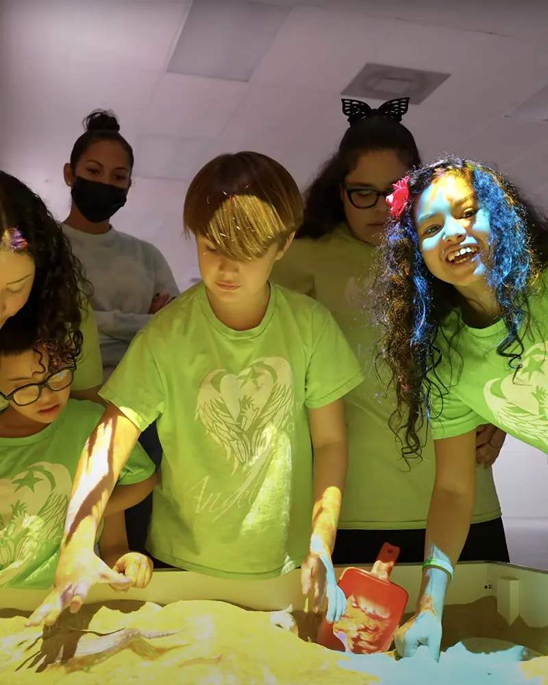 Children in green shirts playing with an interactive illuminated sand table under colorful lights