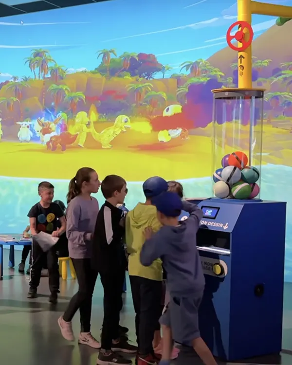 Children interacting with a blue machine in front of a large colorful dinosaur projection