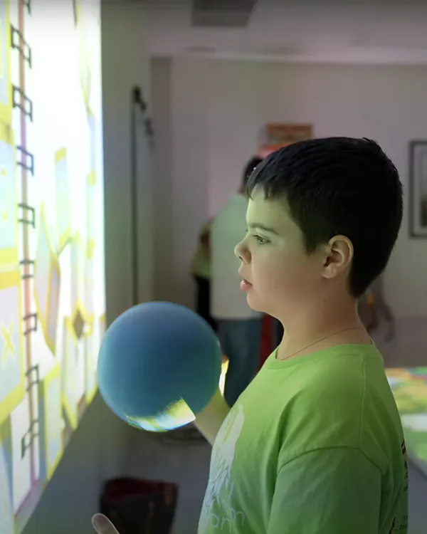 Boy in green shirt holding blue ball while viewing interactive wall projection